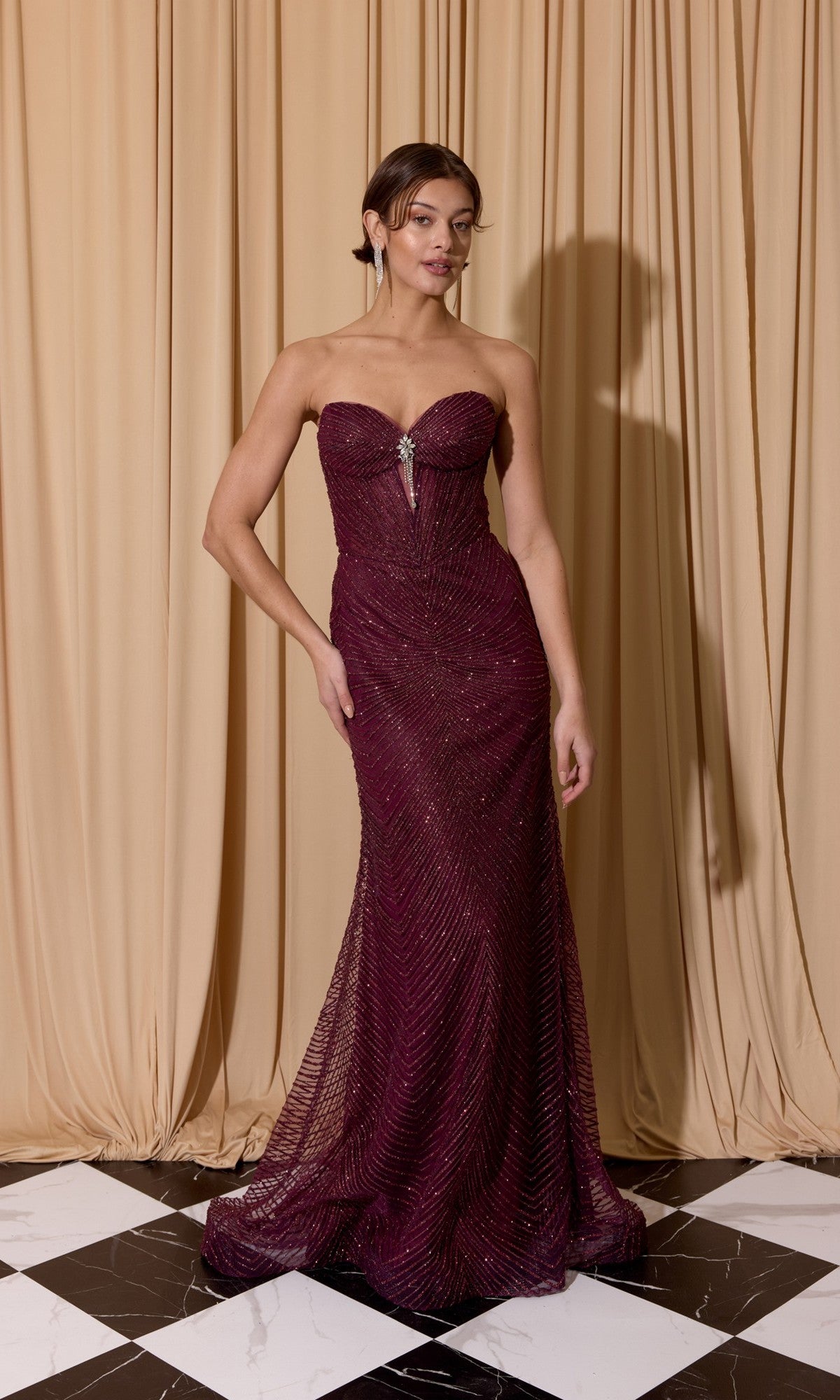 A woman stands on a black and white checkered floor, elegantly wearing the Eureka EK195 Long Prom Dress with a strapless corset bodice and beaded embellishments, in front of beige curtains.